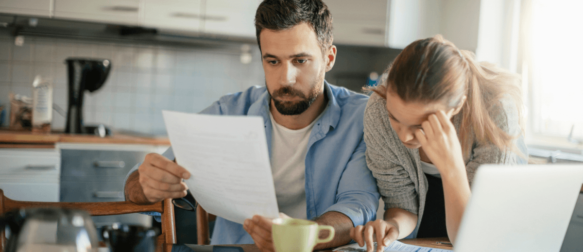A man and woman are sitting at a kitchen table holding financial documents, and they have expressions of stress on their faces.