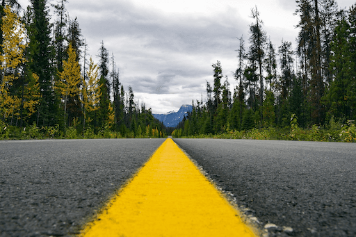 A paved road with a solid yellow line in the center leading toward mountains in the distance.