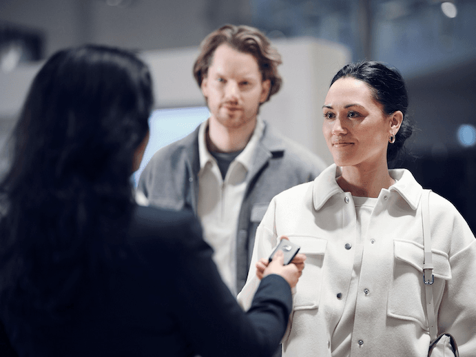 A woman hands keys to another woman who is wearing a white jacket, while a man stands in the background.