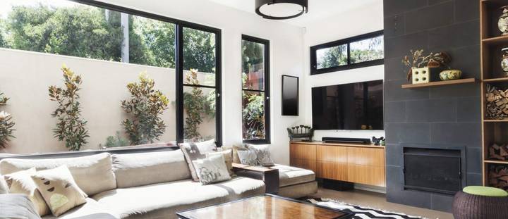 A modern, well-furnished living room with large windows, a stylish fireplace, and lush greenery outside.