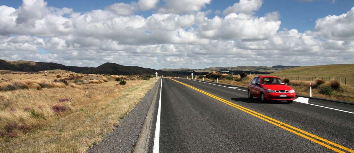 A red car driving on a scenic New Zealand highway illustrating ways to find cheap car insurance with Quashed in 2026.