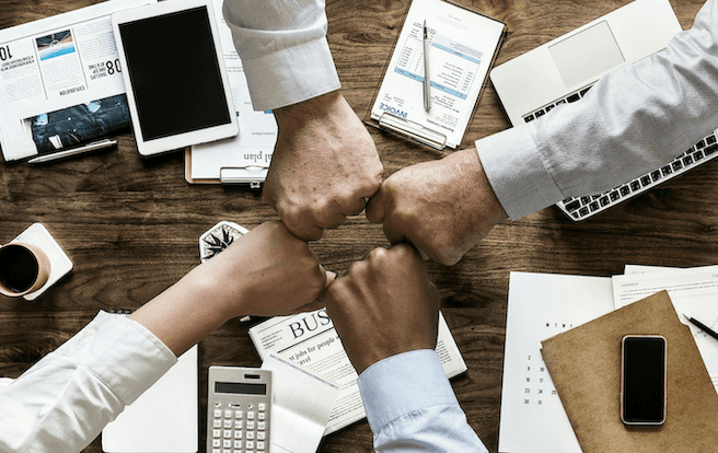 Four people engaging in a fist bump overtop of insurance and financial documents on a crowded desk.