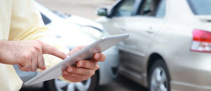 A person holding a tablet while standing in front of two silver cars that have been in a minor collision, demonstrating the importance of having comprehensive car insurance in 2026 via Quashed.