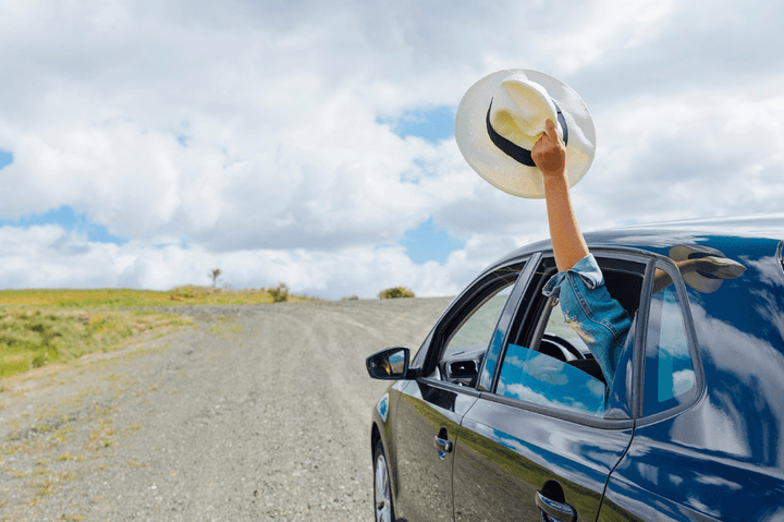 Person waving a hat from a car window on a gravel road. 
