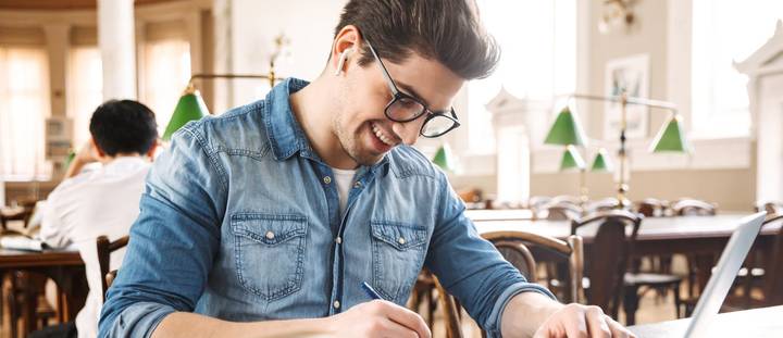 A young male taking notes while working on a laptop.