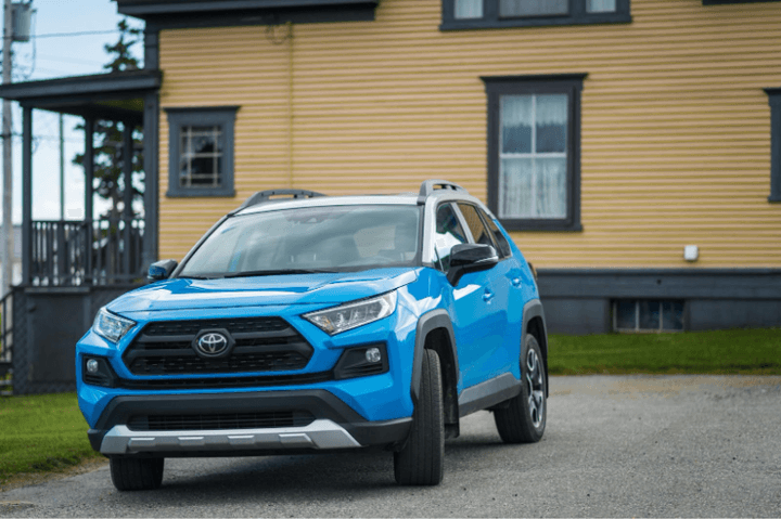 A bright blue Toyota RAV4 with a white two-tone roof parked on a gravel driveway in front of a yellow house with dark trim.